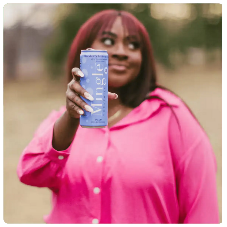 Woman in pink shirt holding a can of Mingle blackberry bellini mocktail outdoors.