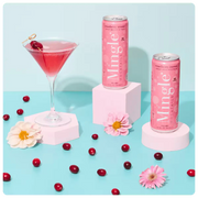 non alcoholic cranberry mocktail being poured into a martini glass, surrounded by cans, cranberries, and flowers on a blue surface