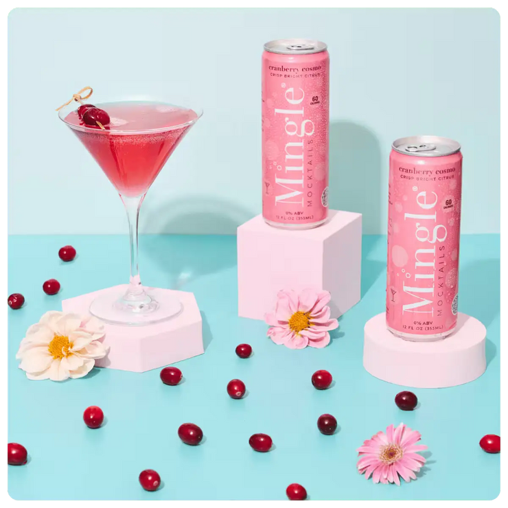 non alcoholic cranberry mocktail being poured into a martini glass, surrounded by cans, cranberries, and flowers on a blue surface