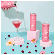 non alcoholic cranberry mocktail being poured into a martini glass, surrounded by cans, cranberries, & flowers on a blue surface