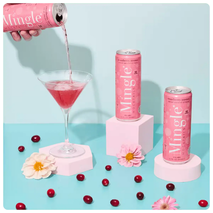 non alcoholic cranberry mocktail being poured into a martini glass, surrounded by cans, cranberries, & flowers on a blue surface