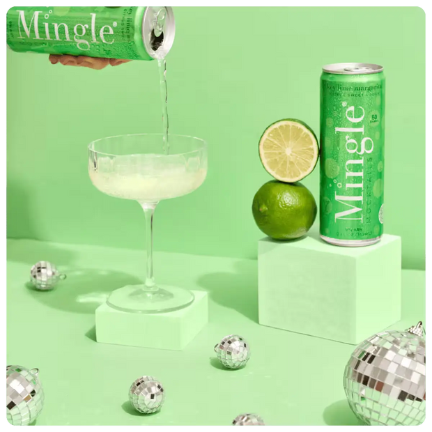 margarita mocktail with lime being poured into a cocktail glass, with a can, lime, and decorative disco balls on a green background