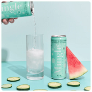 non alcoholic cucumber mojito cocktail being poured into a glass, with cucumber slices, and a watermelon slice on a blue background