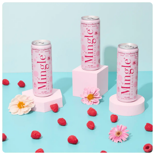 raspberry rosé mocktails being poured into a glass, with cans, fresh raspberries, and flowers arranged on a blue surface