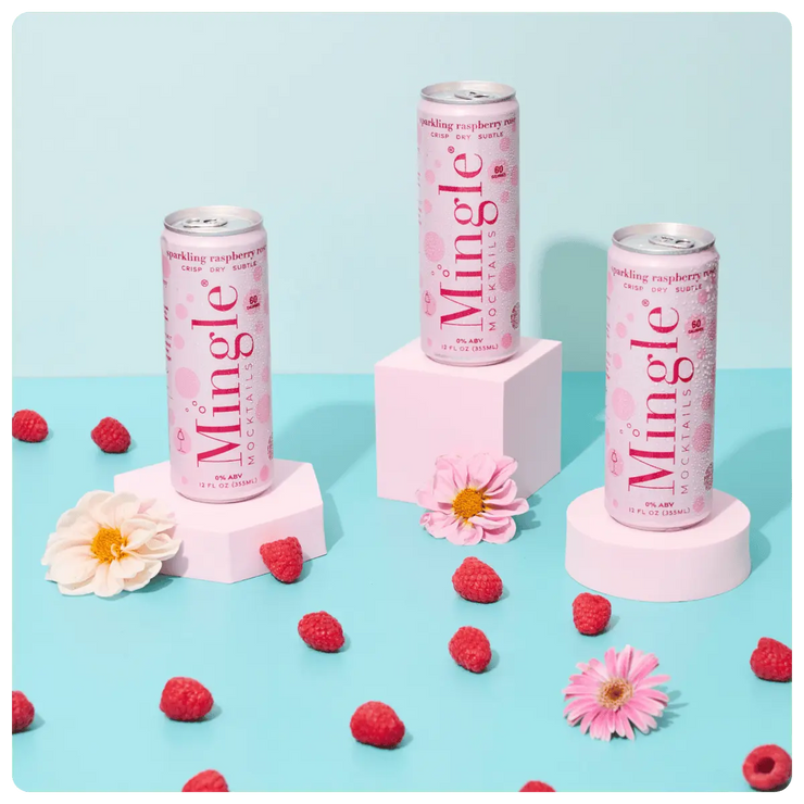 raspberry rosé mocktails being poured into a glass, with cans, fresh raspberries, and flowers arranged on a blue surface